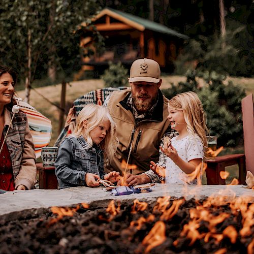 A family sits around a campfire in a forested area near a cabin, enjoying an outdoor gathering together.