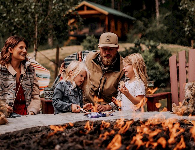 A family sits around a campfire in a forested area near a cabin, enjoying an outdoor gathering together.