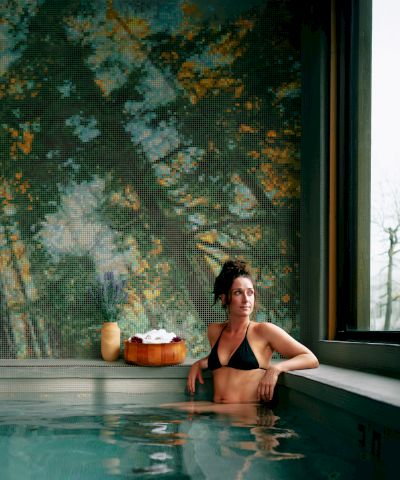 A person sits in a tranquil indoor pool, relaxing by the edge with a drink, a peaceful forest mural on the wall behind, and large windows letting in light.