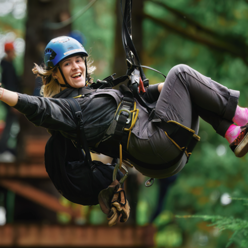 A person smiling and giving a thumbs-up while zip-lining through a forest, wearing a helmet and harness for safety.
