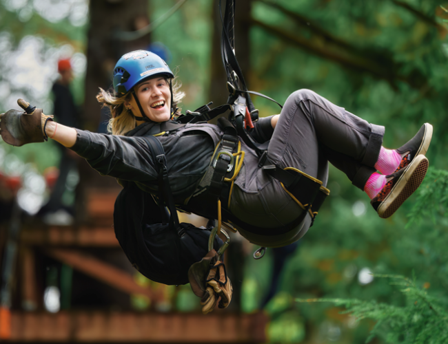 A person smiling and giving a thumbs-up while zip-lining through a forest, wearing a helmet and harness for safety.