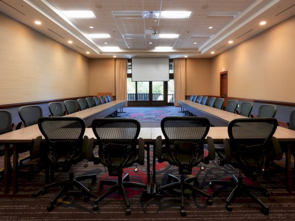 The image shows a conference room with a U-shaped table arrangement, multiple chairs, a large screen, and overhead lights.