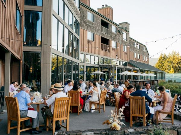 People are dining outdoors at a restaurant with a modern wooden exterior, enjoying a pleasant setting under string lights.