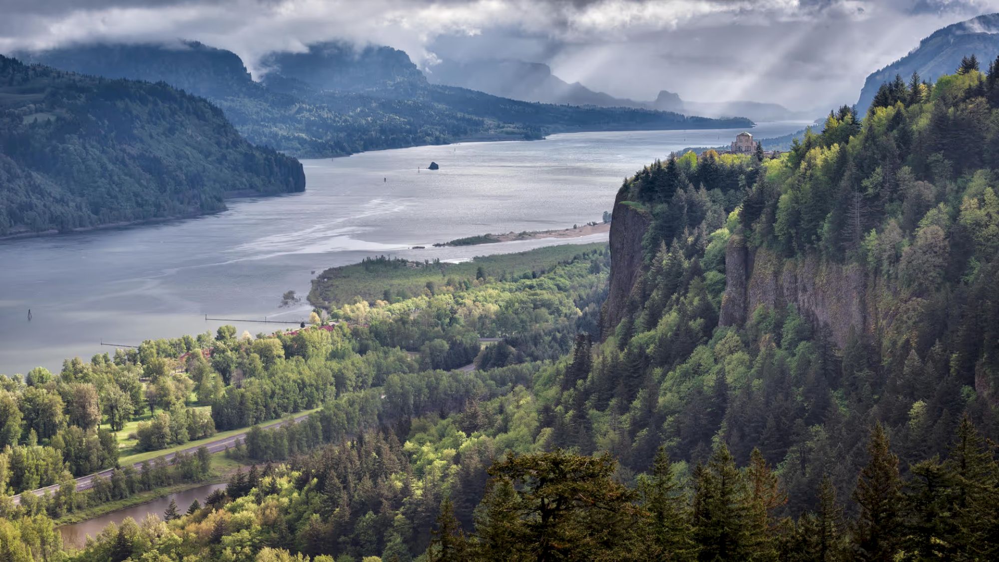 A scenic landscape with a river winding through lush green hills and forests, beneath a cloudy sky, with rays of sunlight piercing through.