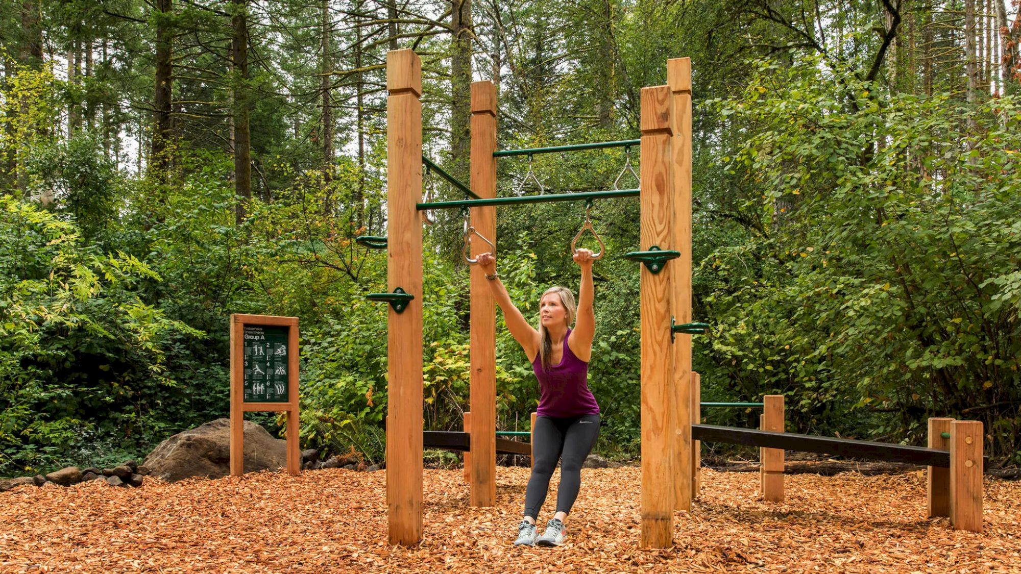A person is exercising on outdoor gym equipment in a scenic forest setting, surrounded by trees and woodchip ground cover.