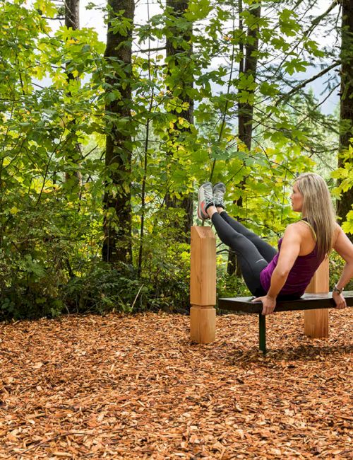 A person exercises on outdoor gym equipment surrounded by trees, with an instruction sign visible nearby.