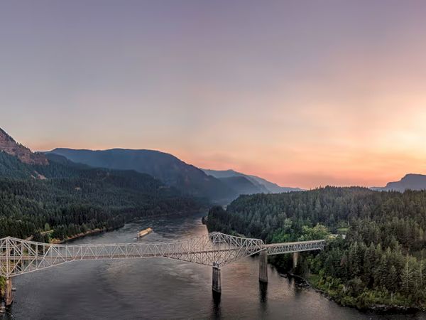 A bridge spans a river, surrounded by forested mountains under a colorful sunset sky.
