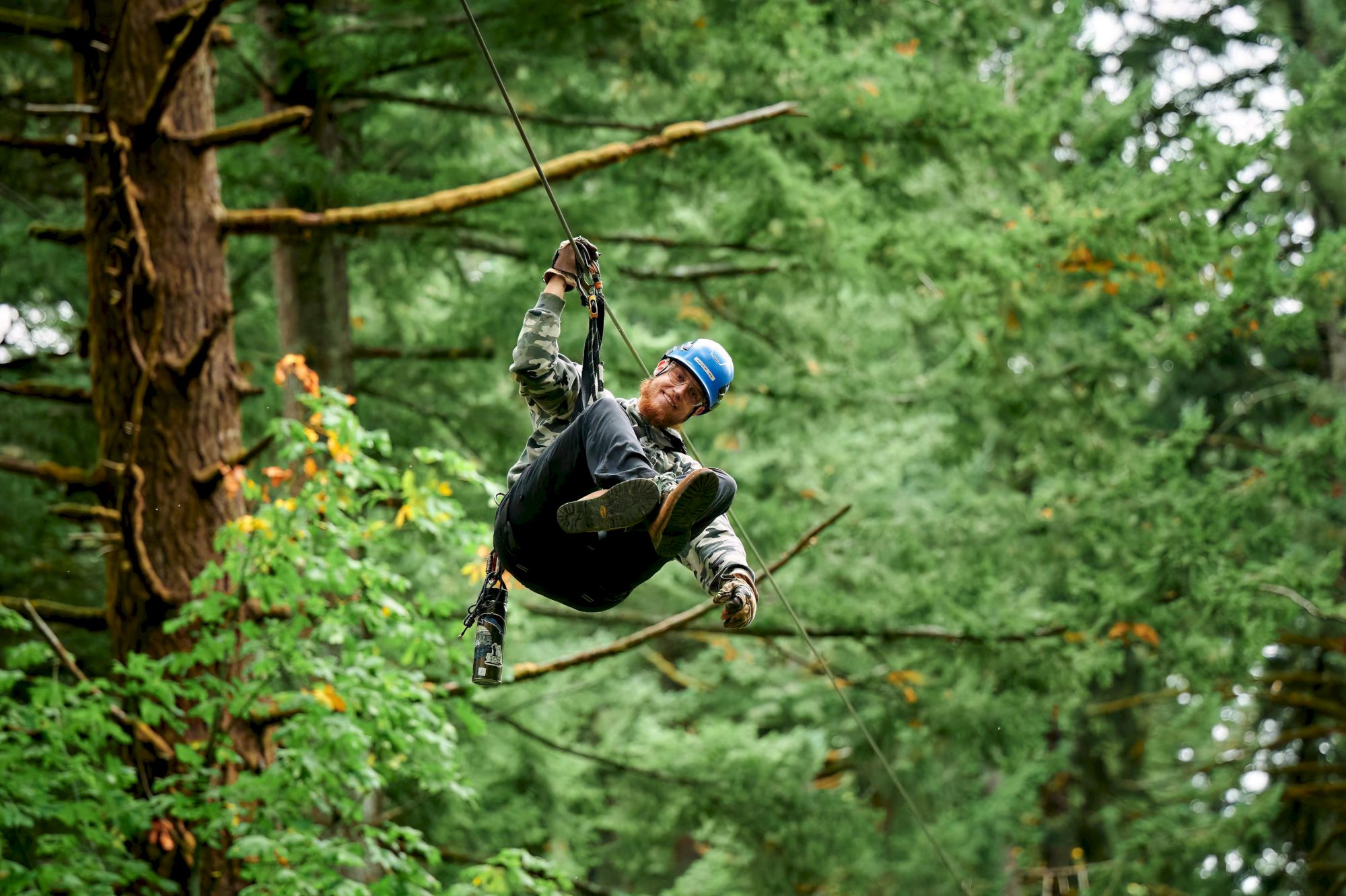 A person in a blue helmet is ziplining through a forest, surrounded by green trees, arms holding the line above.