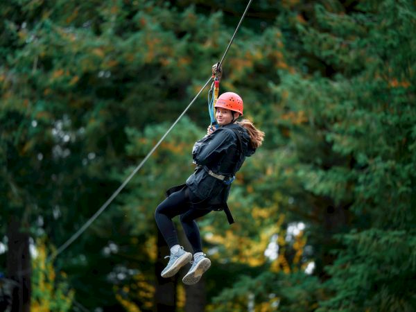 A person wearing a helmet and harness is zip-lining through a forested area, surrounded by green trees.