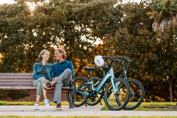 A couple sits on a park bench, chatting and smiling, with two bicycles and a helmet parked nearby, against a backdrop of trees.