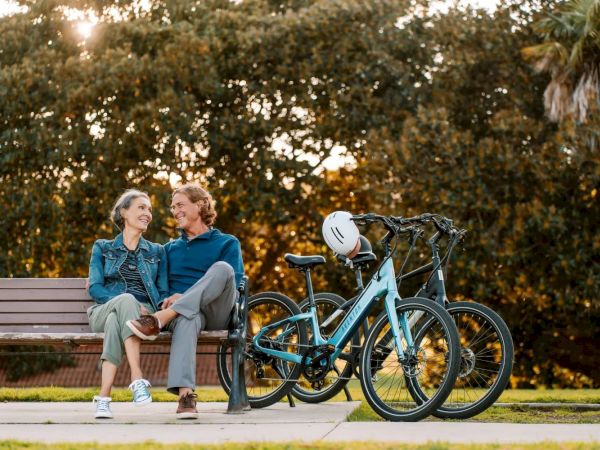 A couple sits on a park bench, chatting and smiling, with two bicycles and a helmet parked nearby, against a backdrop of trees.