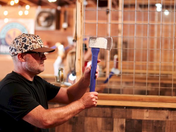 A person in a camo hat, holding an axe in an indoor axe-throwing venue, ready to throw.