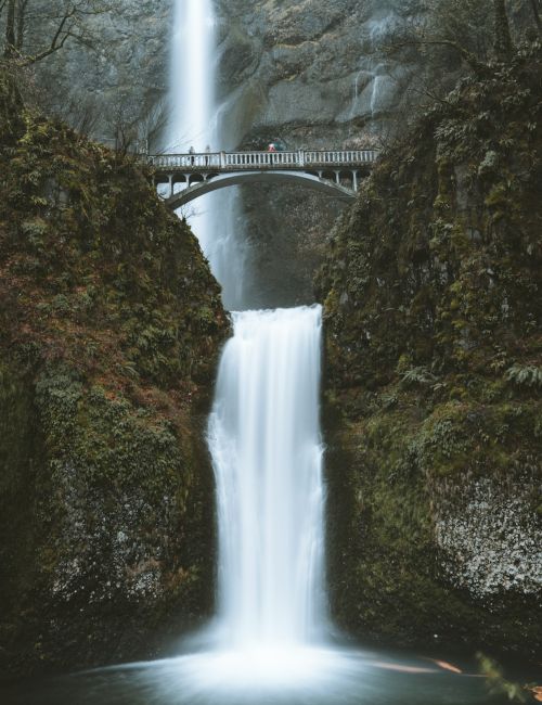 A waterfall cascades down with a bridge crossing above, surrounded by lush greenery and rocky cliffs, creating a serene natural scene.