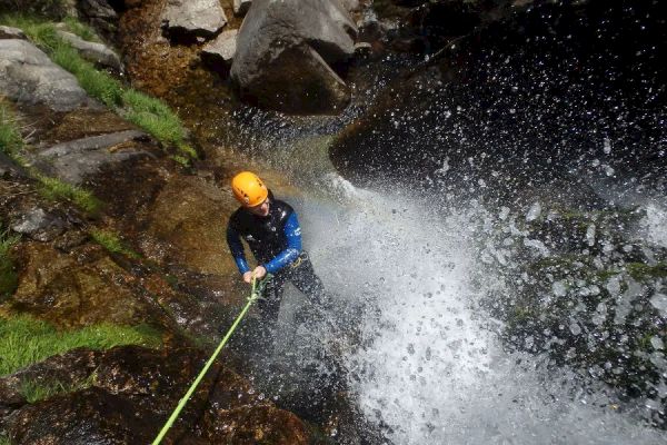 A person in a helmet is rappelling down a waterfall, surrounded by rocks and vegetation, holding onto a rope amidst splashing water.