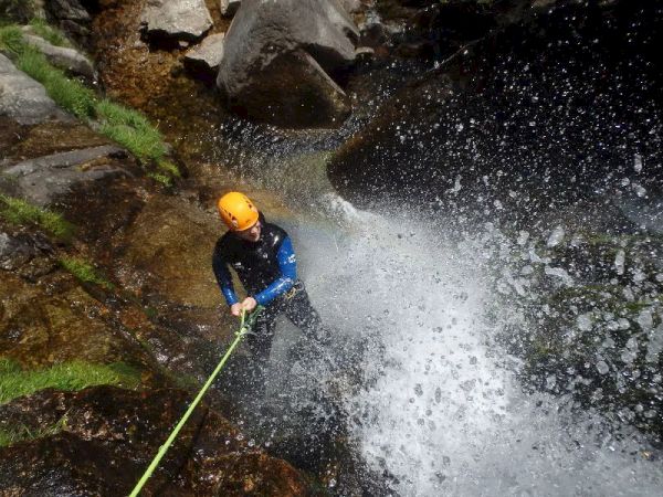A person in a helmet is rappelling down a waterfall, surrounded by rocks and vegetation, holding onto a rope amidst splashing water.