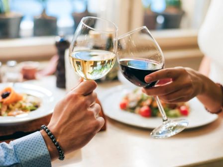 Two people clinking glasses of white and red wine at a table set with plates of food, creating a celebratory atmosphere.