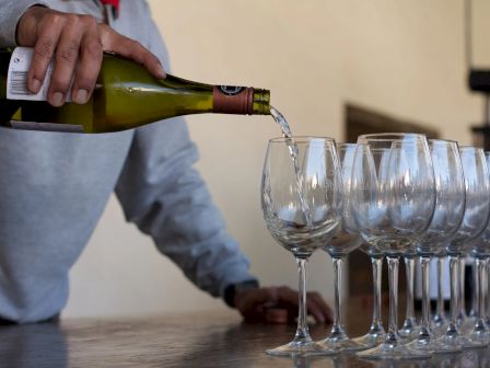 A person is pouring white wine into a row of clear wine glasses lined up on a surface.