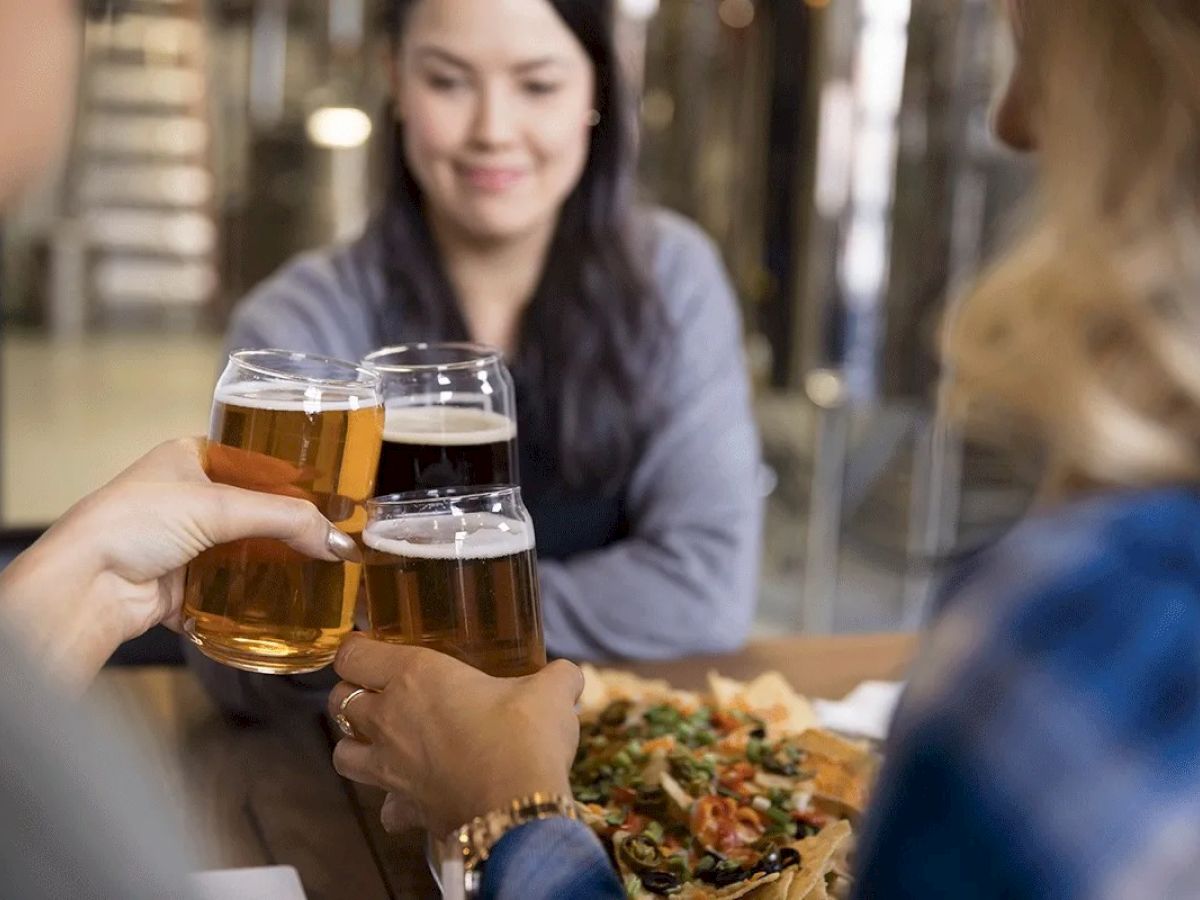 Three people clinking glasses filled with beer, with a plate of food on the table, and a woman smiling in the background.