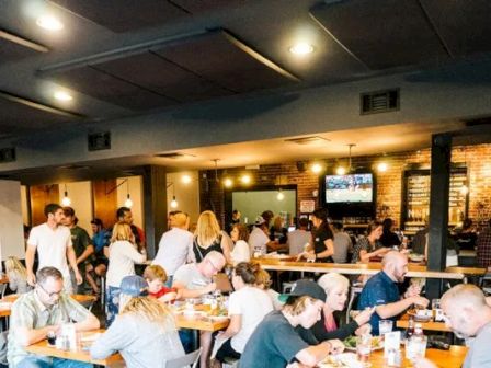 A busy restaurant scene with people dining and socializing at tables, under warm lighting with bar, decor, and televisions visible.
