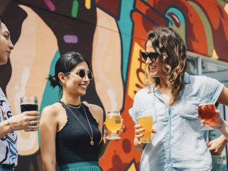 Three people smiling and holding drinks in front of a colorful mural, enjoying a sunny day outdoors.