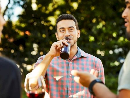 A man in a checkered shirt drinks from a bottle, chatting with two others holding a glass and gesturing, in a sunny outdoor setting.