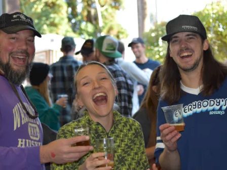 Three people are laughing and enjoying drinks at a social gathering, with others in the background.