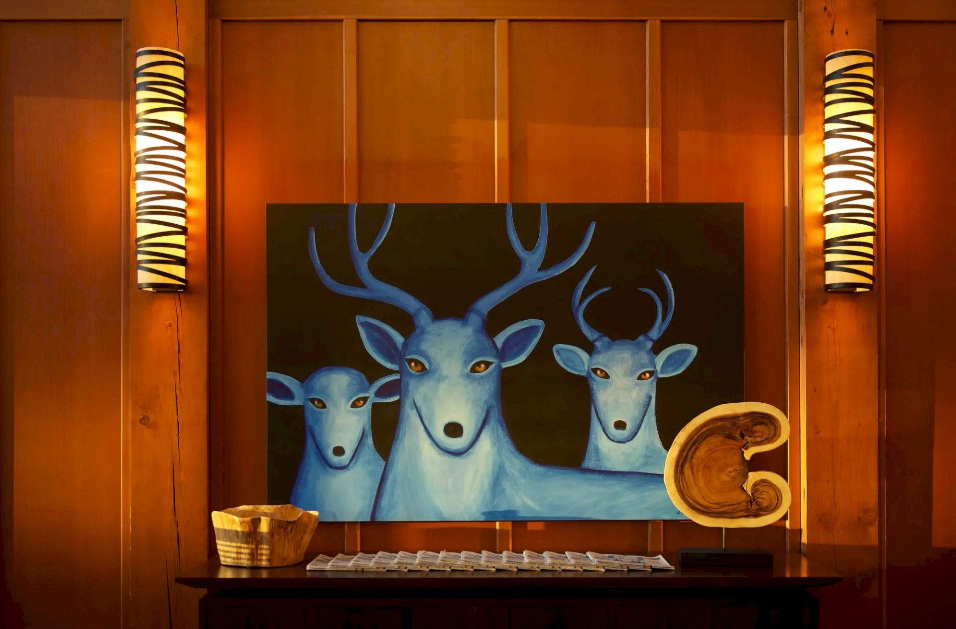 The image shows a painting of three blue deer displayed on a wooden wall, flanked by two tall lamps and decorative items on a shelf.