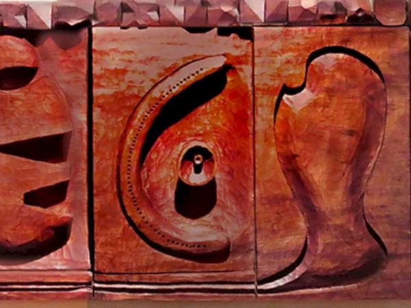 The image shows a wooden sculpture featuring abstract shapes and carved human faces, arranged in a series of panels.