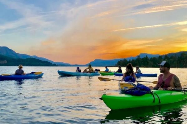 People are kayaking on a calm lake during sunset, surrounded by mountains and a colorful sky.