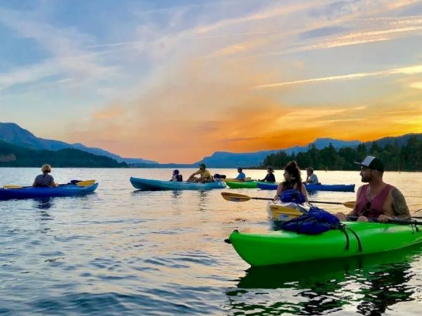 People are kayaking on a calm lake during sunset, surrounded by mountains and a colorful sky.