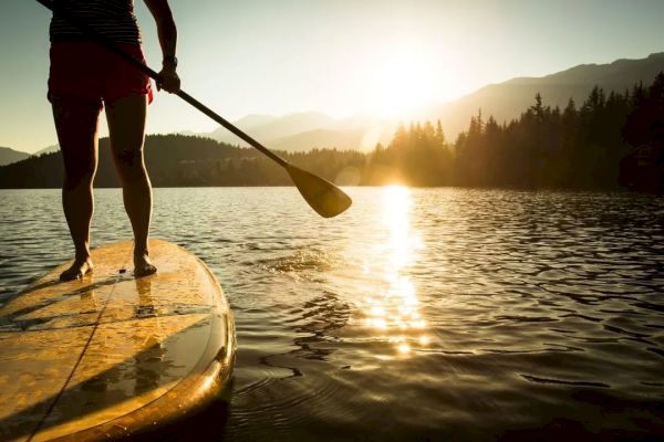 A person is paddleboarding on a lake during sunset, with mountains and trees in the background, creating a serene atmosphere.
