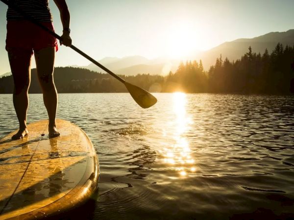 A person is paddleboarding on a lake during sunset, with mountains and trees in the background, creating a serene atmosphere.