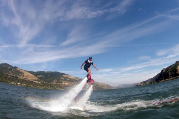 Two people are enjoying a flyboarding experience over a body of water, with mountains in the background under a clear sky.