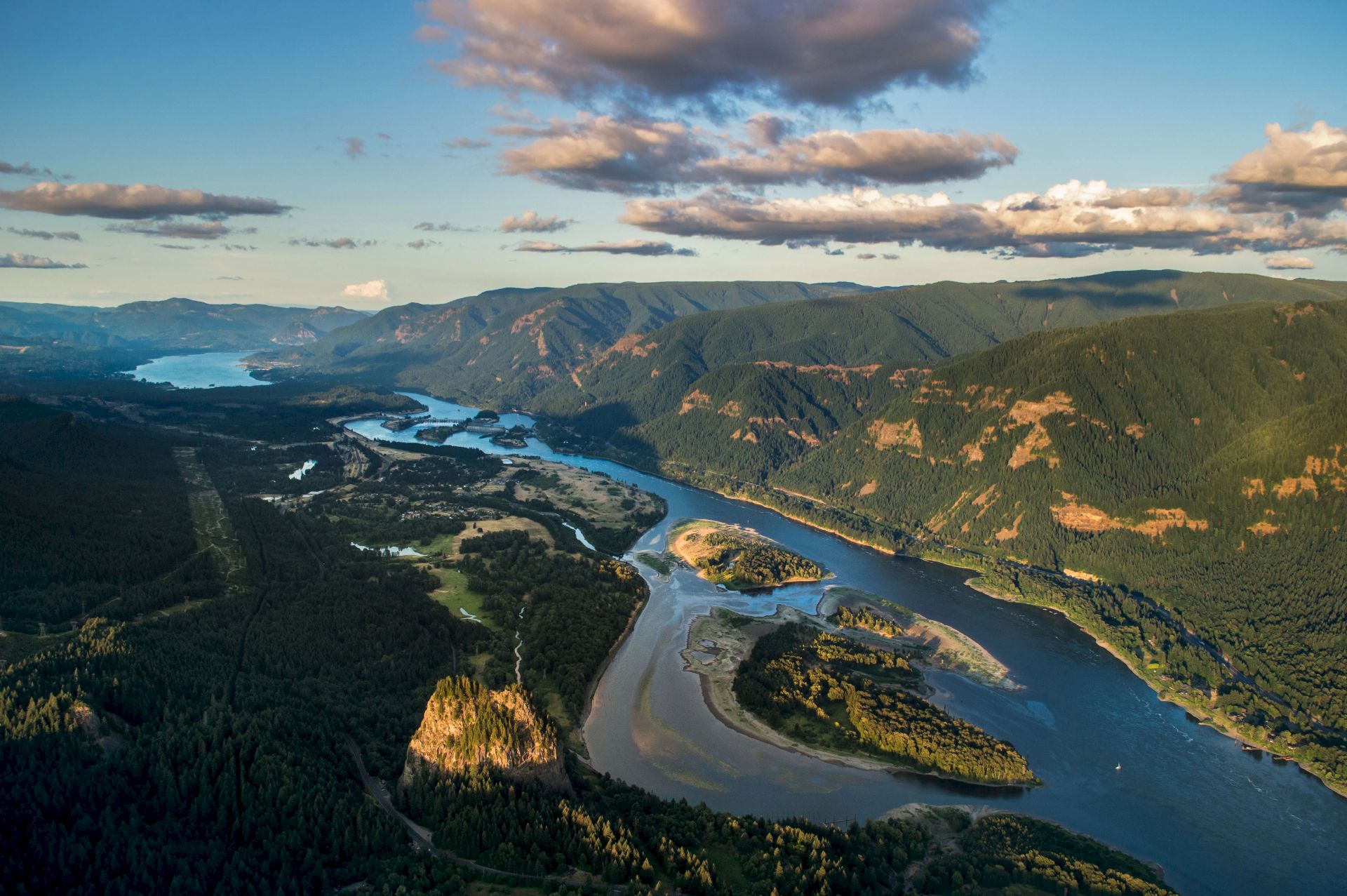 An aerial view of a winding river cutting through lush, forested mountains under a sky with clouds at sunset.