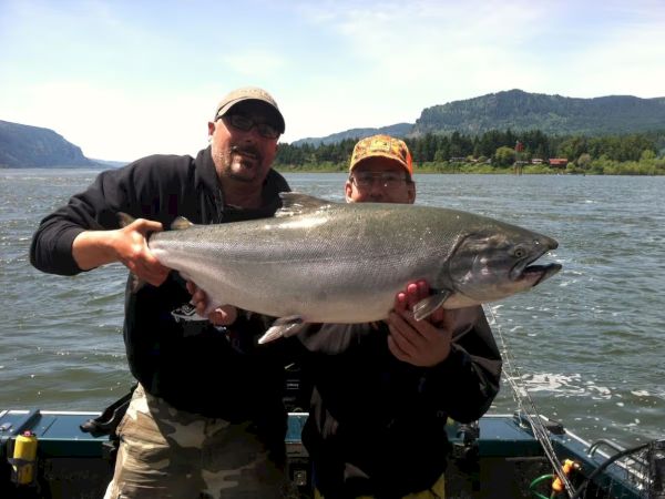 Two people are on a boat holding a large fish, with a scenic lake and mountains in the background.