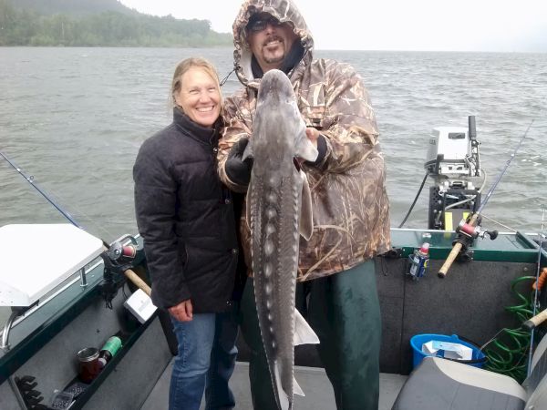 A man and woman on a boat, holding a large fish and smiling. It's a cloudy day on the water, and they seem content.