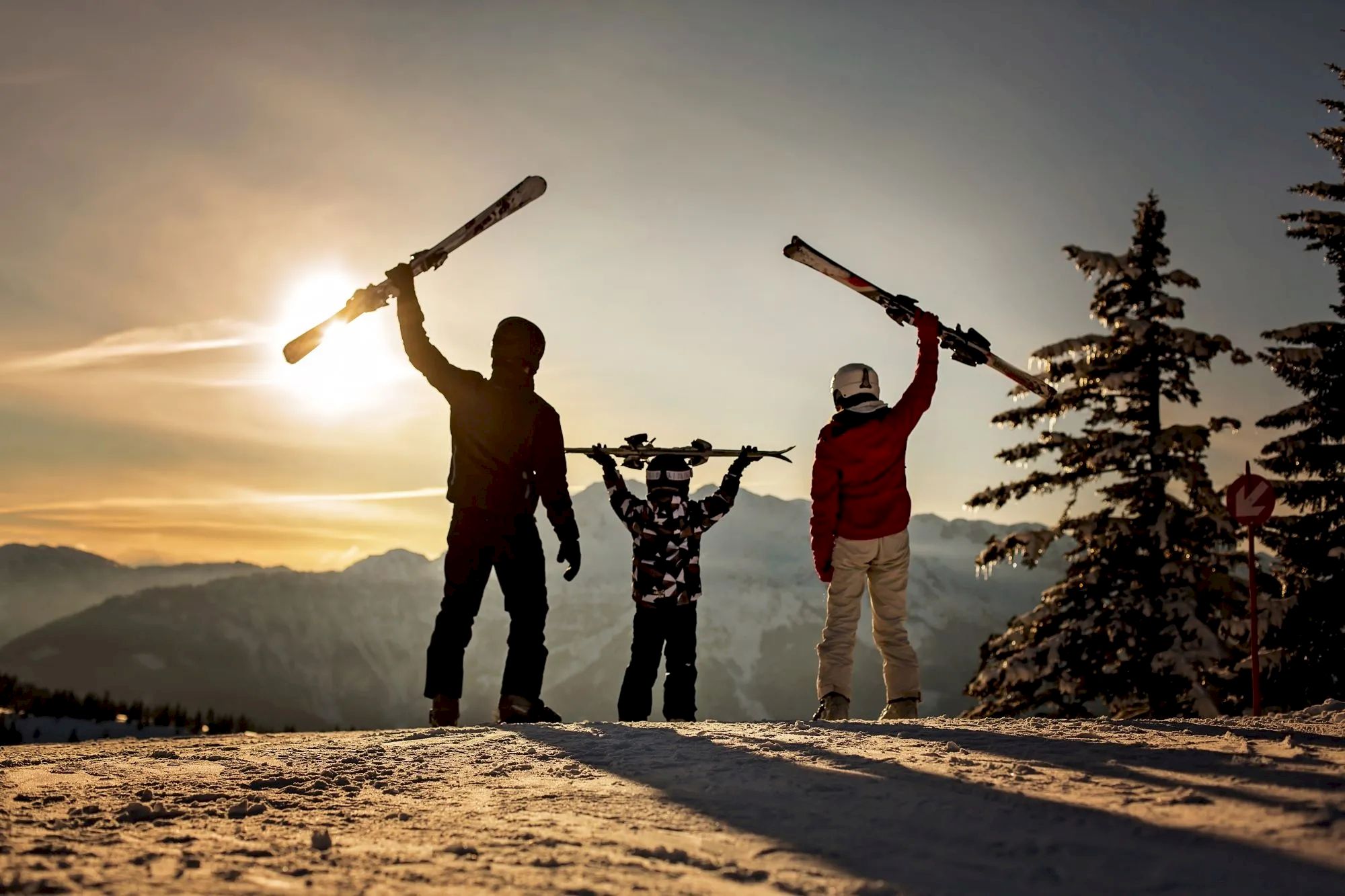 Three people holding skis aloft on a snowy mountain at sunset, with trees and mountains in the background.