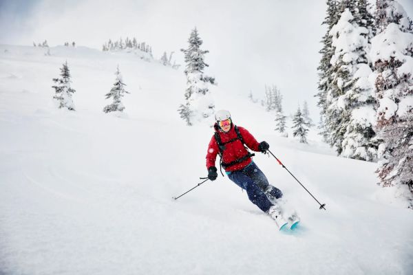 A person is skiing down a snowy slope surrounded by snow-covered trees, wearing a red jacket and helmet.