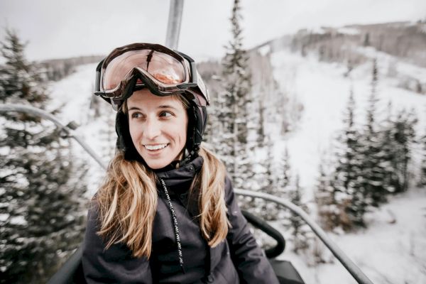 A person wearing ski gear and goggles is sitting on a ski lift surrounded by snowy trees and slopes, smiling in the winter setting.