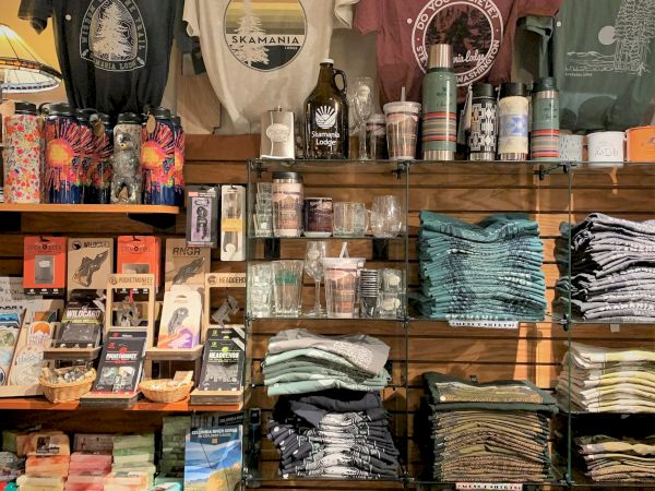 This image shows a retail display with T-shirts, water bottles, postcards, books, candles, and stacks of towels or clothes on shelves.