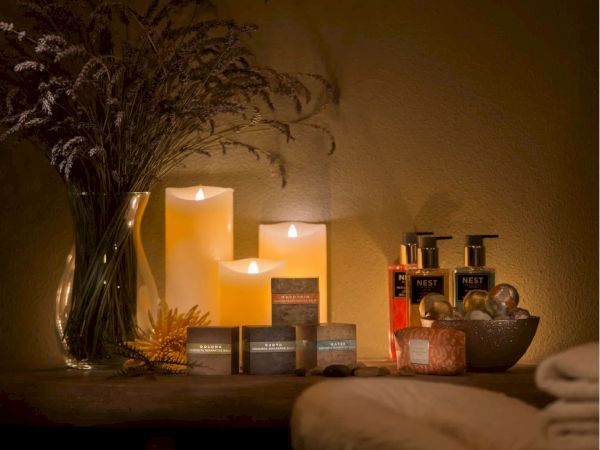 The image shows a cozy spa setting with lit candles, lotions, aromatherapy soaps, a vase of dried flowers, and folded towels on a table.
