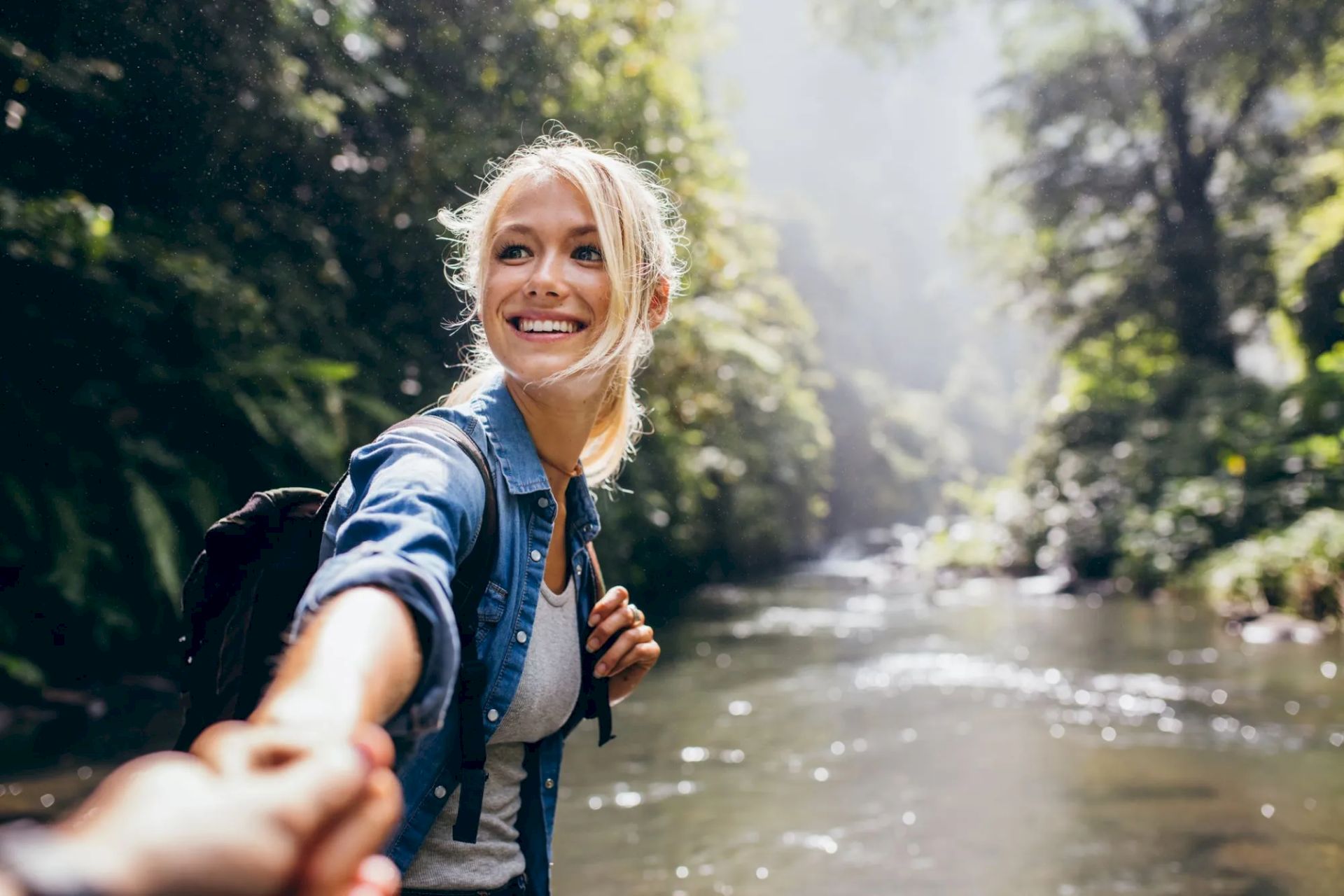 A person in a denim shirt is smiling, holding someone's hand near a flowing river surrounded by lush greenery.