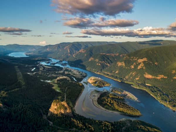 A winding river cuts through a lush, mountainous landscape under a partly cloudy sky, with dense green forests on either side.