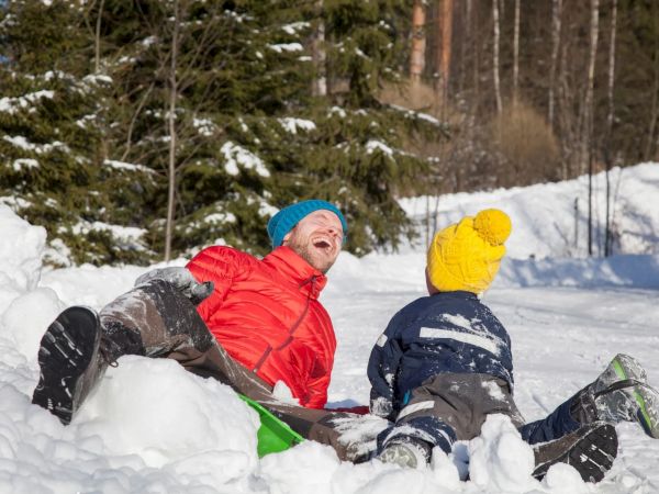 Two people are sitting and laughing in the snow, surrounded by trees, wearing colorful winter clothing.