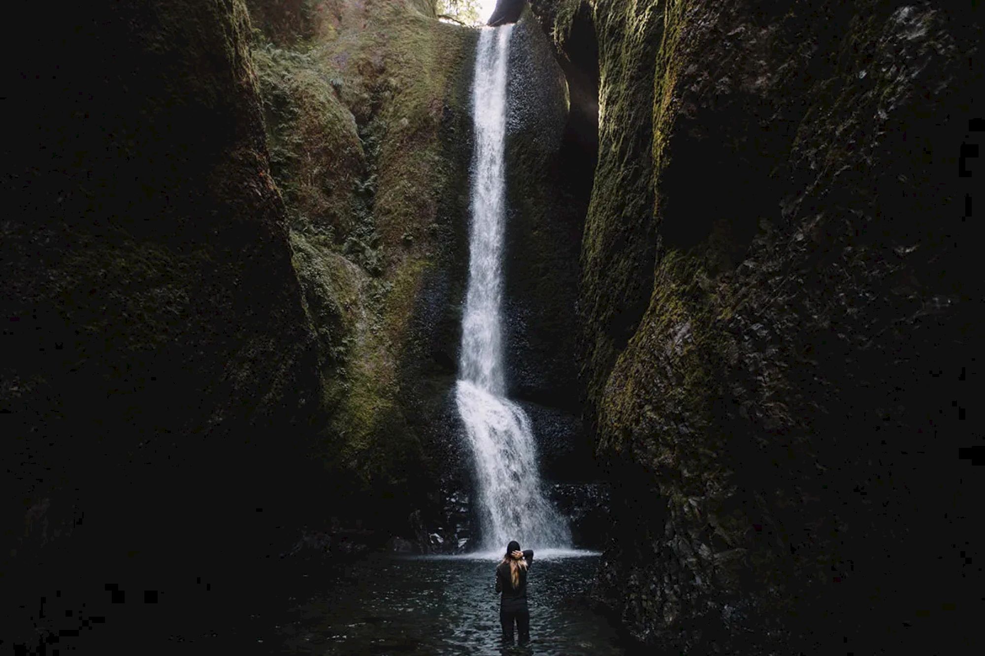 A waterfall cascades down a rocky cliff surrounded by moss, with a person standing in the water below, capturing the serene scene.