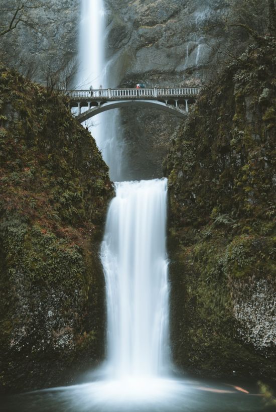 A scenic waterfall with a stone bridge above, surrounded by lush green cliffs and trees, creating a serene natural setting.
