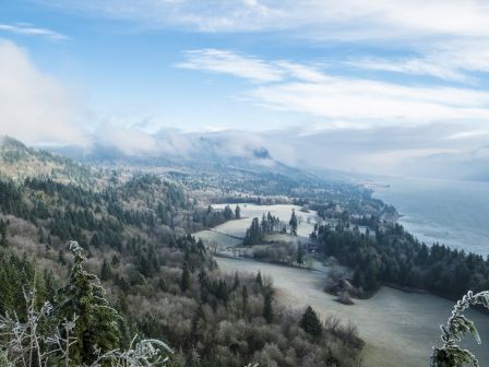 A snowy hillside overlooks a coastline with evergreen trees, misty clouds, and a distant town by the water, stretching toward the horizon.