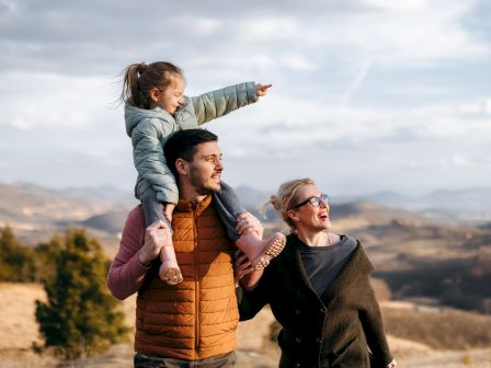 A family of three enjoys a scenic view in a hilly landscape; the child on the man's shoulders points into the distance as they all smile.