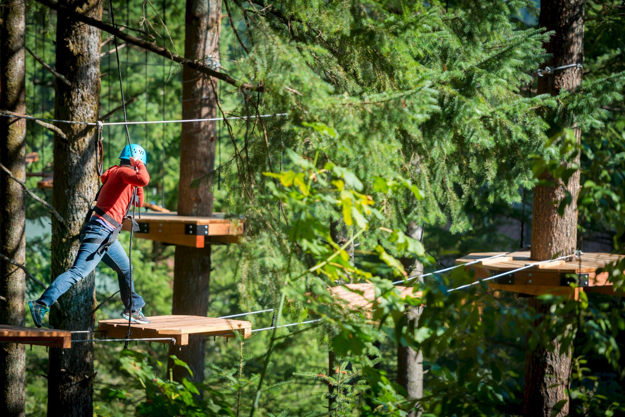 A person in outdoor gear navigates a treetop ropes course, balancing on wooden platforms and wearing a helmet in a forest setting.