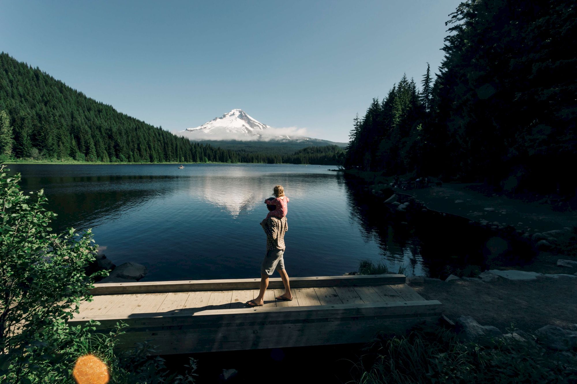A person carries a child on a wooden bridge by a lake, with a snow-covered mountain and dense forest in the background, under a clear sky.